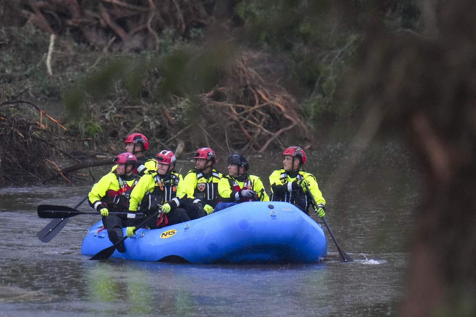 Six rescue workers wearing protective gear paddle an inflatable boat down a river.