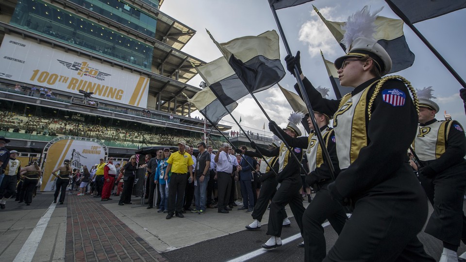 Students in black and gold marching-band uniforms hold Purdue flags and walk in tandem. 