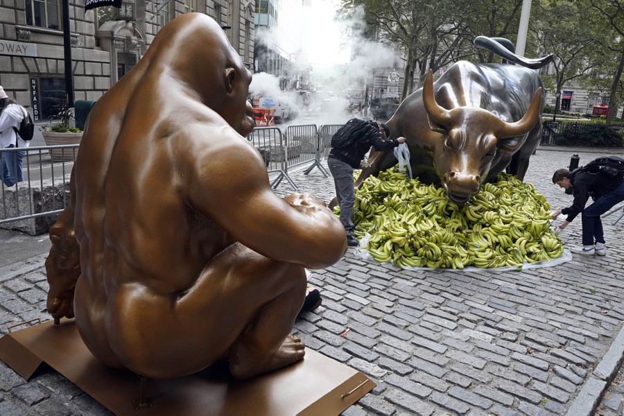 A large bronze-colored statue of a seated gorilla faces a large statue of a charging bull surrounded by a pile of bananas.