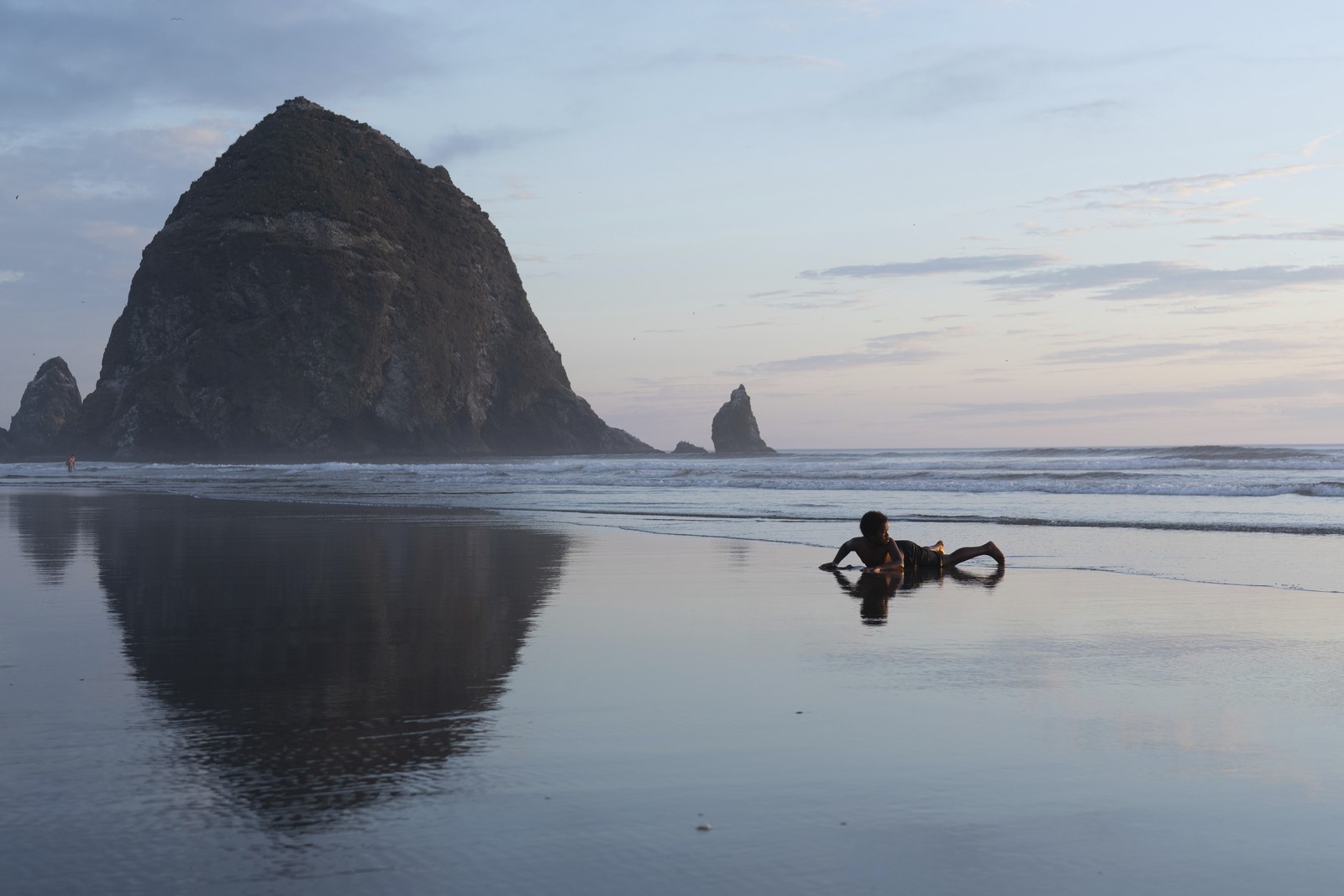 A boy lays down on a beach, with a tall rock formation visible in the background.