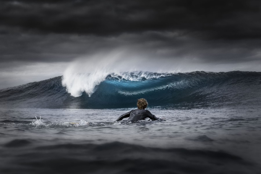 A surfer paddles toward a breaking wave under a dark, cloudy sky.
