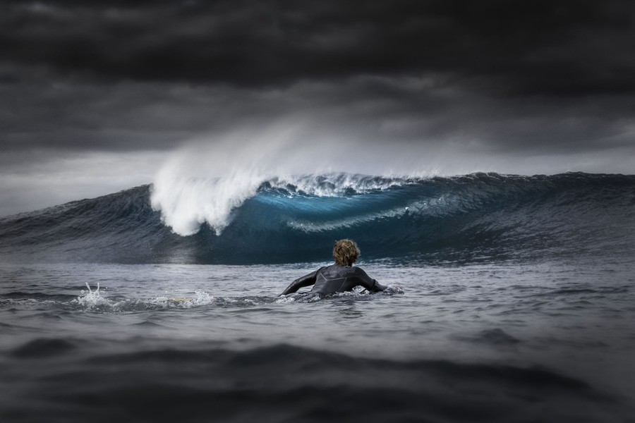Un surfista rema hacia una ola rompiendo bajo un cielo oscuro y nublado.