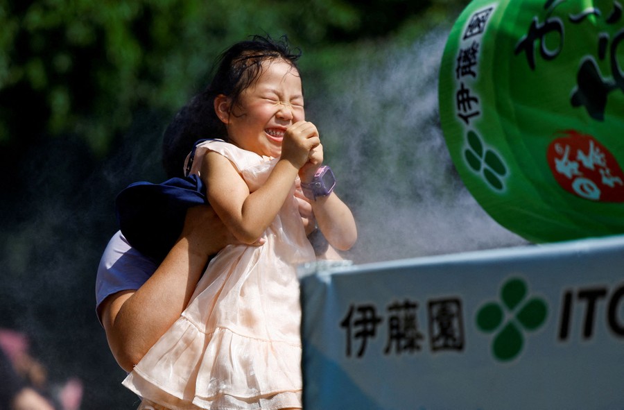 A child holds her hands together and scrunches her eyes shut, grinning, as someone holds her up in front of a cooling mister.
