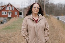 A woman wearing a brown jacket stands on the side of the road in front of a red house