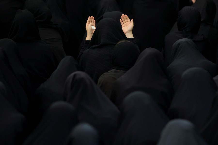 More than a dozen people photographed from behind, all wearing dark head coverings, with one at the center holding her hands up in prayer