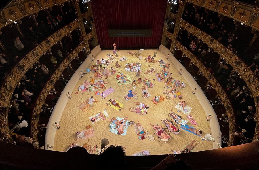 An audience stands inside an older theatre, looking down at a large floor below, where performers in swimsuits rehearse on a stage covered with beach sand.