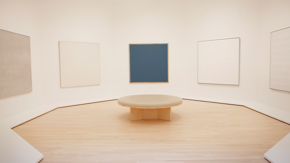 a white room with Agnes Martin's paintings on the wall and a round seat in the center