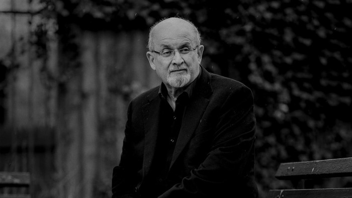 A black-and-white photograph of Salman Rushdie sitting on a park bench.