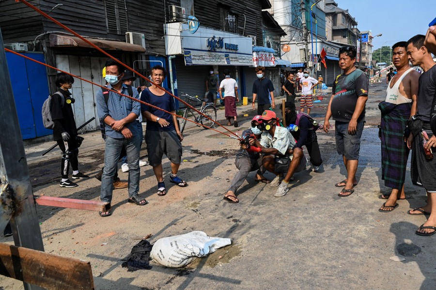 People stand in a street and watch as several others pull back on a long elastic cord, testing a slingshot.