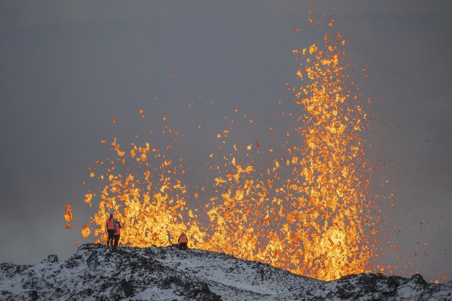 Several people stand on a ridge in front of jets of erupting lava.