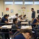 Students take a test in a classroom; a teacher stands at the front of the room. 