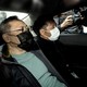 Three men sit in the backseat of a car with masks on as photographers stand outside.