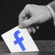 A hand puts a ballot into a ballot box. The ballot has the Facebook logo illustrated on it.