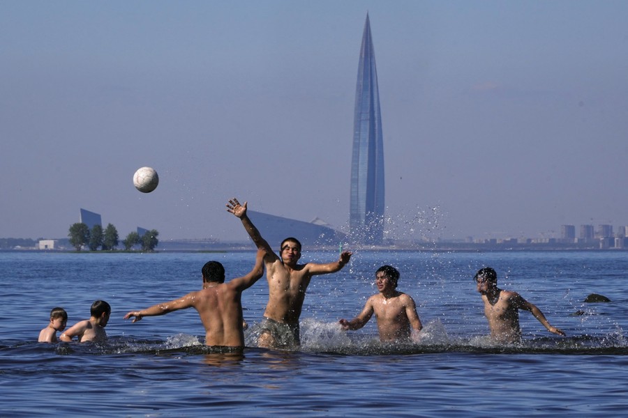Several people play volleyball in waist-deep water, with a lone skyscraper visible in the distance.