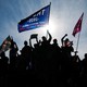 Trump voters raise their fists and wave pro-Trump flags in a protest. A blue sky is seen behind them.