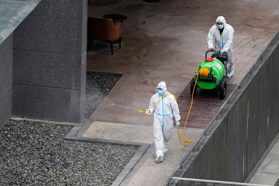 Workers in protective suits spray disinfectant in a courtyard.