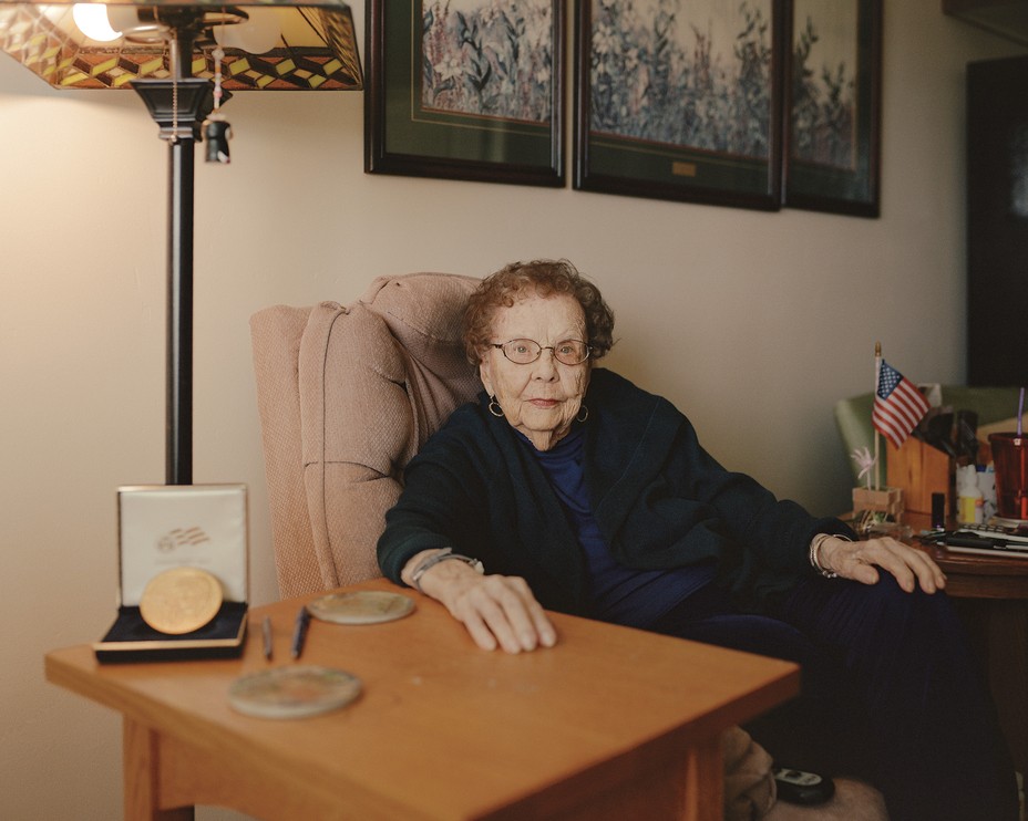 color photo of woman with short red hair and glasses sitting in armchair with medal and small American flag on tables at her side