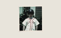 A cream colored background has a photo in the center of a little boy, maybe 5 years old. He has fair skin and is wearing a dark-blue Boston Redsocks baseball helmet and white jersey with red trim that says "Red Sox." Behind him in the background is a shrub and the siding on a house.