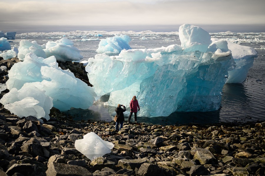 Two people take photographs on a rocky shoreline beside large icebergs.