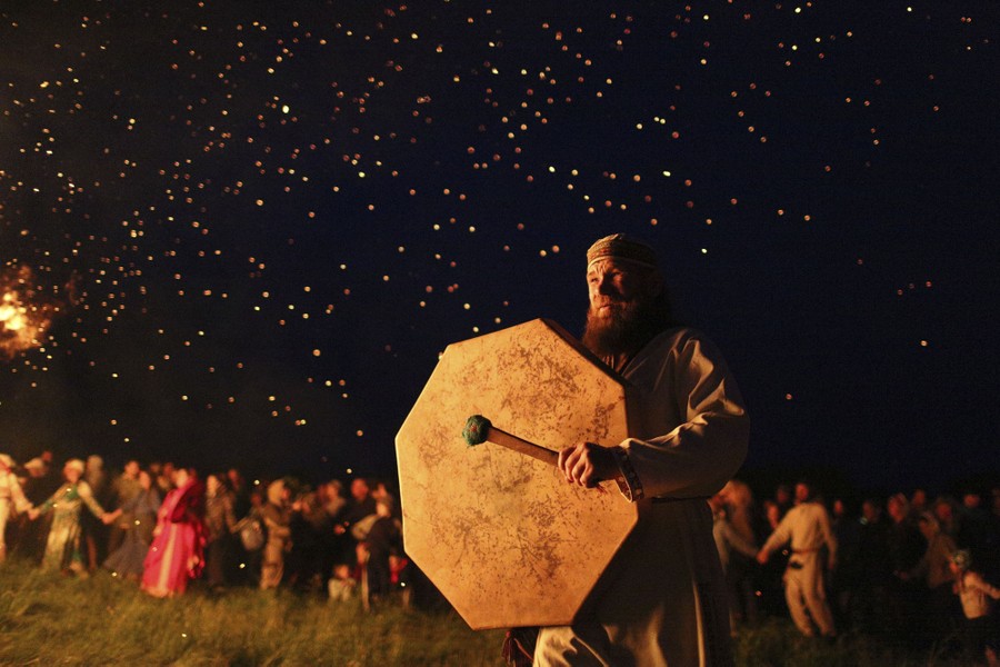 A man beats a drum, in front of a crowd, near a bonfire.