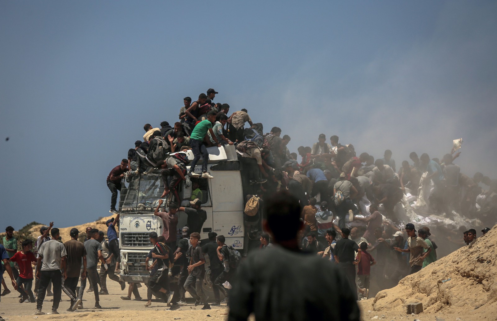 Dozens of people climb onto the cab and bed of a flatbed delivery truck in a chaotic scene.