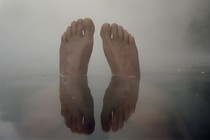 A pair of feet coming out of the water, against a foggy grey sky, and reflected in the water