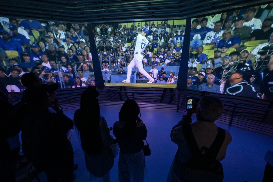 Several visitors stand in an exhibition space looking toward three large video screens depicting a baseball game.