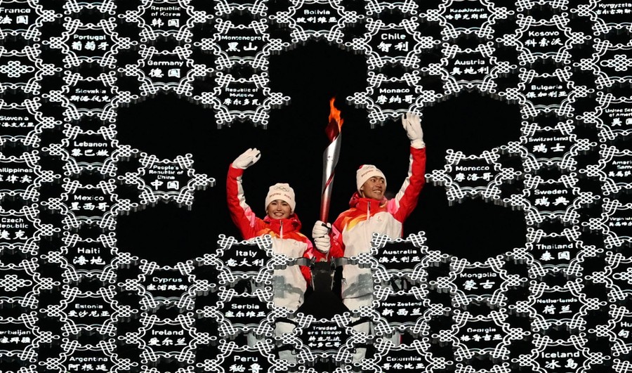 Two people wave while holding an Olympic torch, framed by a sculptural snowflake with the names of participating countries.