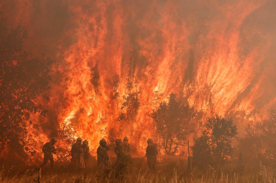 About eight firefighters stand together in a field in front of a huge wildfire.