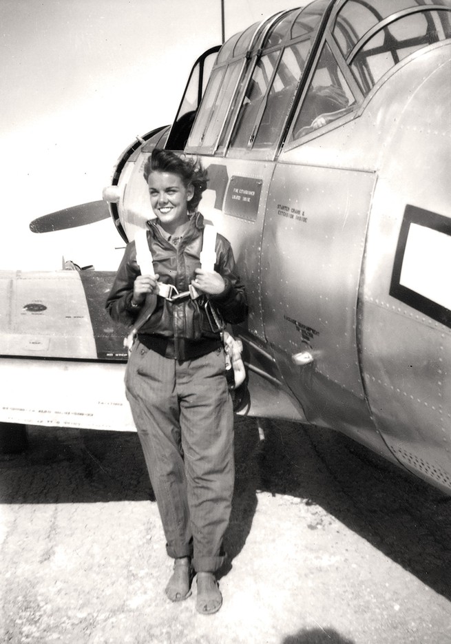 black-and-white archival photo of smiling woman in flight gear, leather jacket, and sandals standing next to her plane