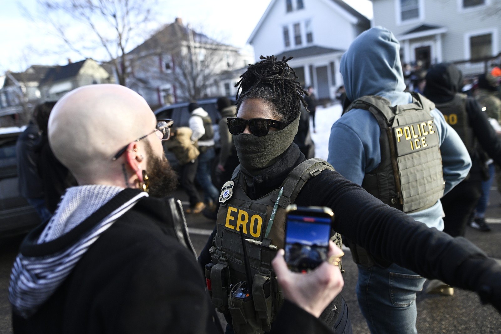A federal agent wearing a mask holds out their arm, trying to hold back civilians who gathered at the scene of an immigration arrest.