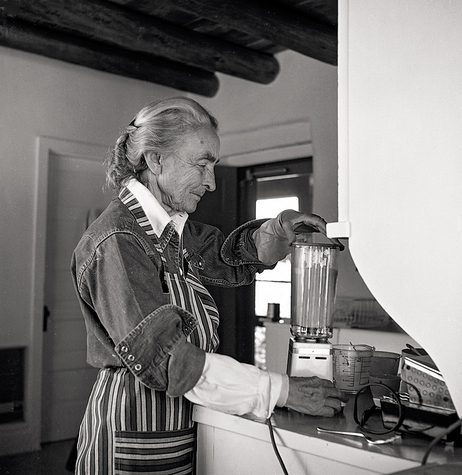 black-and-white photo of O'Keeffe wearing white collared shirt, denim jacket, and striped apron standing at kitchen counter using a blender