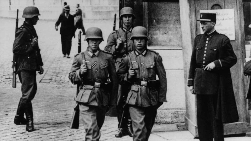 Black-and-white photo of four Nazi soldiers walking through a city street with rifles strapped to their backs as a policeman watches