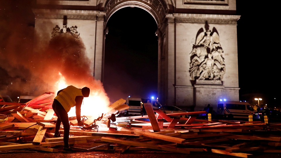 A protester wearing a yellow vest burns a barricade in front of a stone arch.