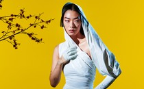 Rina Sawayama wearing a white dress with a hood against a yellow background