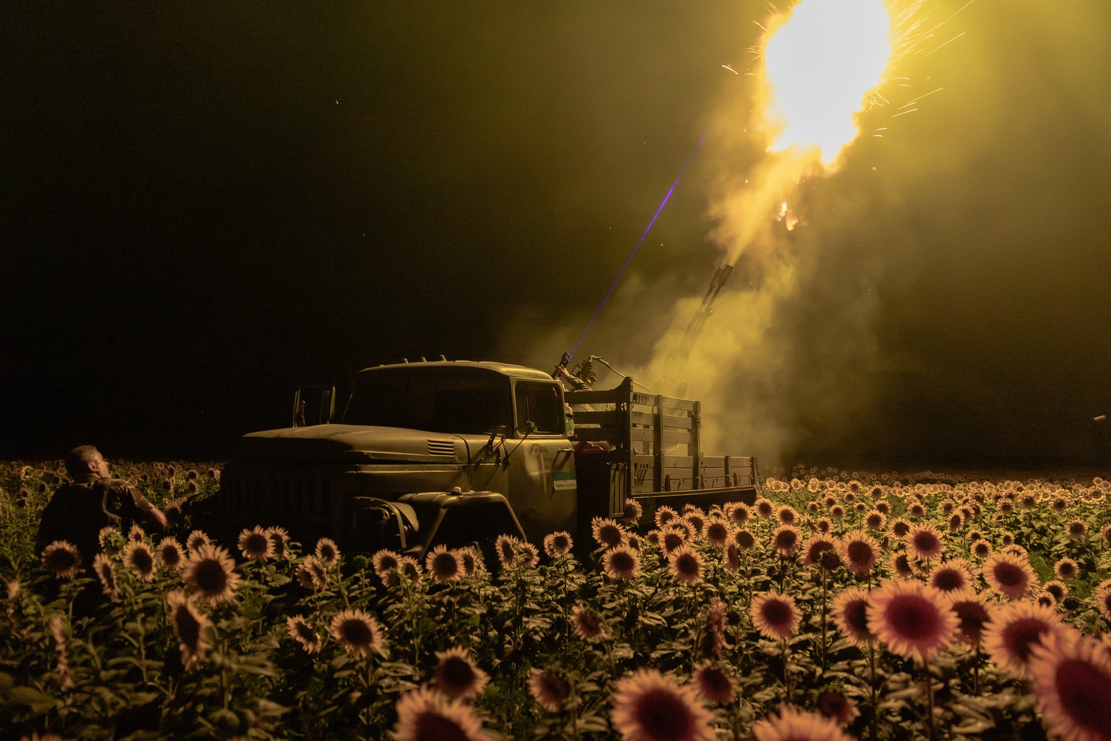 Soldiers fire a large anti-aircraft gun from the back of a truck in a sunflower field, at night, the muzzle flash illuminating the sunflowers.