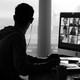 A black-and-white photo of a man working at a desk in his apartment. A Zoom meeting is open on his computer screen, and he seems to be looking out a window.