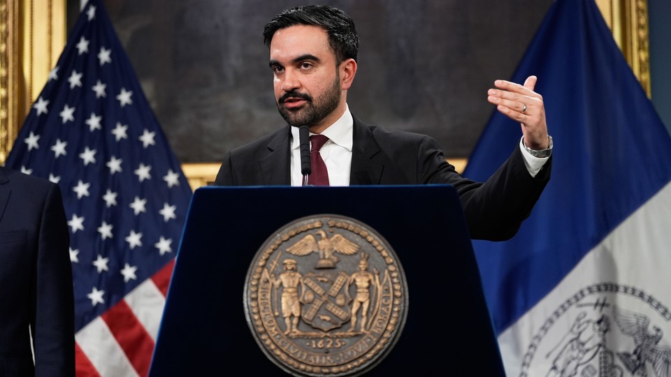 A photo of Mayor Zohran Mamdani speaking at a podium in front of an American flag and a New York City flag.