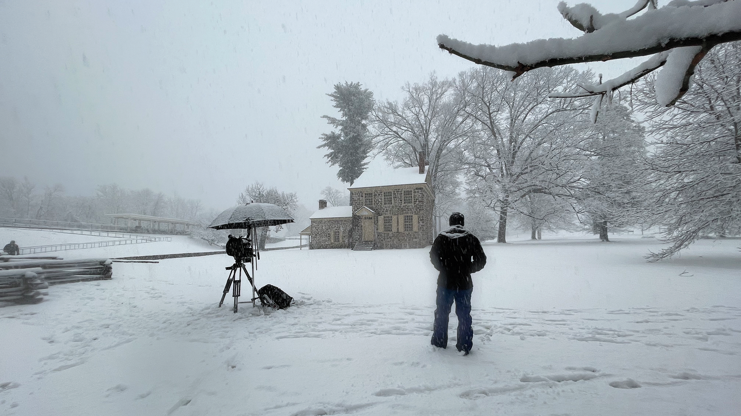 e camera on tripod covered with umbrella in snow-covered landscape with colonial house and split-rail fence