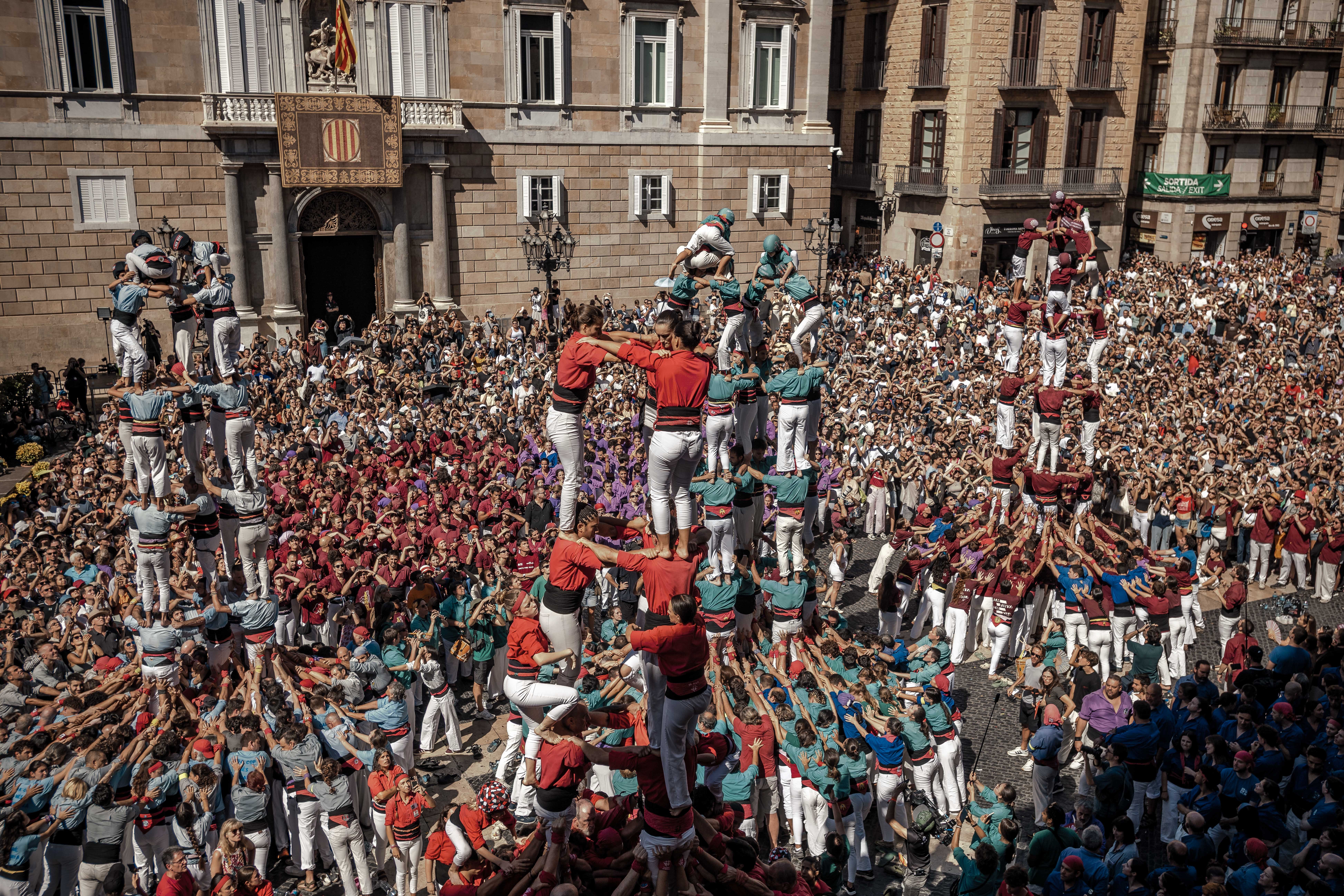 At least four different teams of people build human towers in a plaza, watched by hundreds standing nearby.