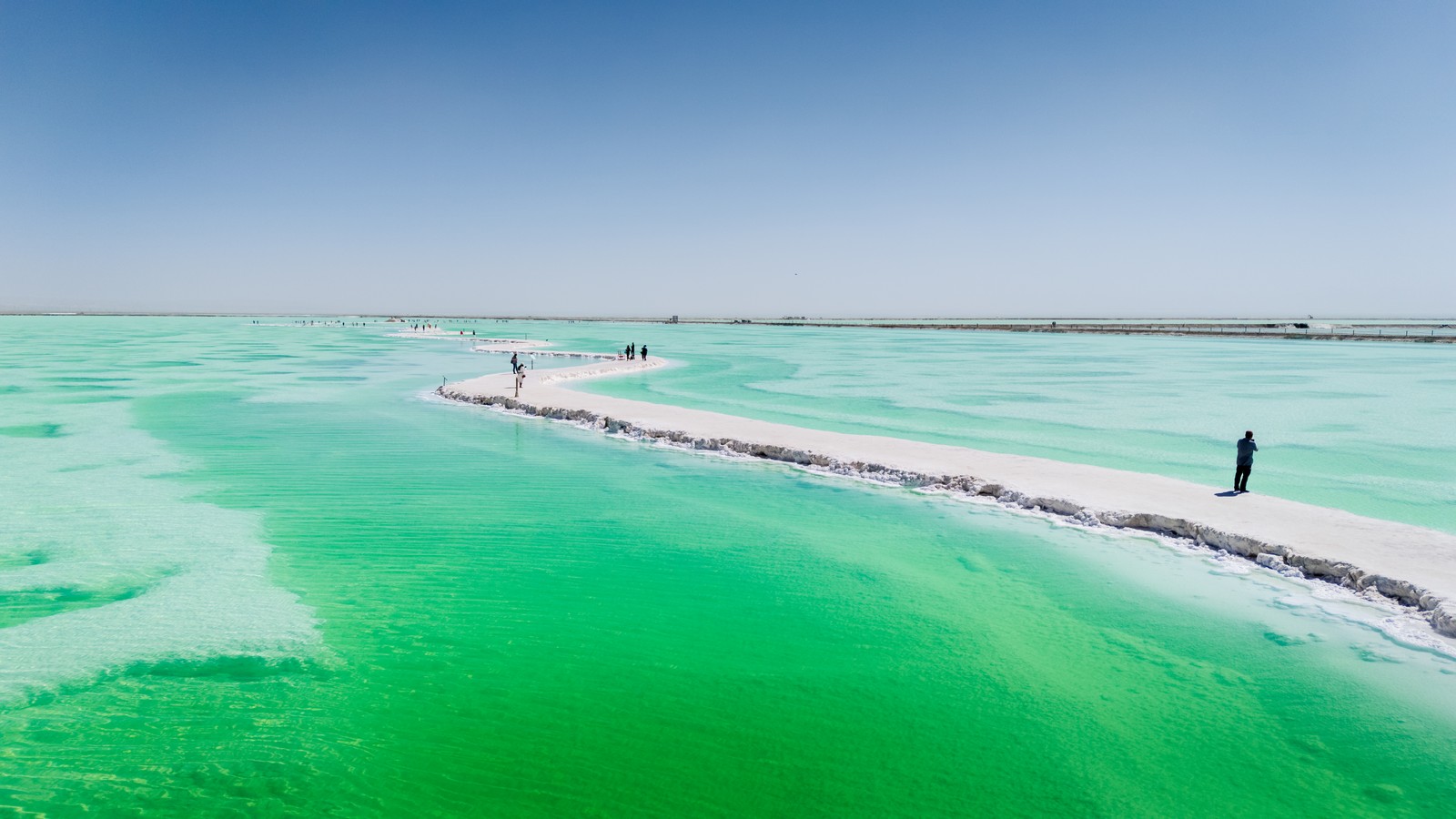 Several people walk along a path through a salt lake filled with bright green water.
