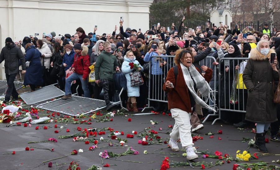 A large crowd walks past toppled barriers and flowers strewn on the ground.