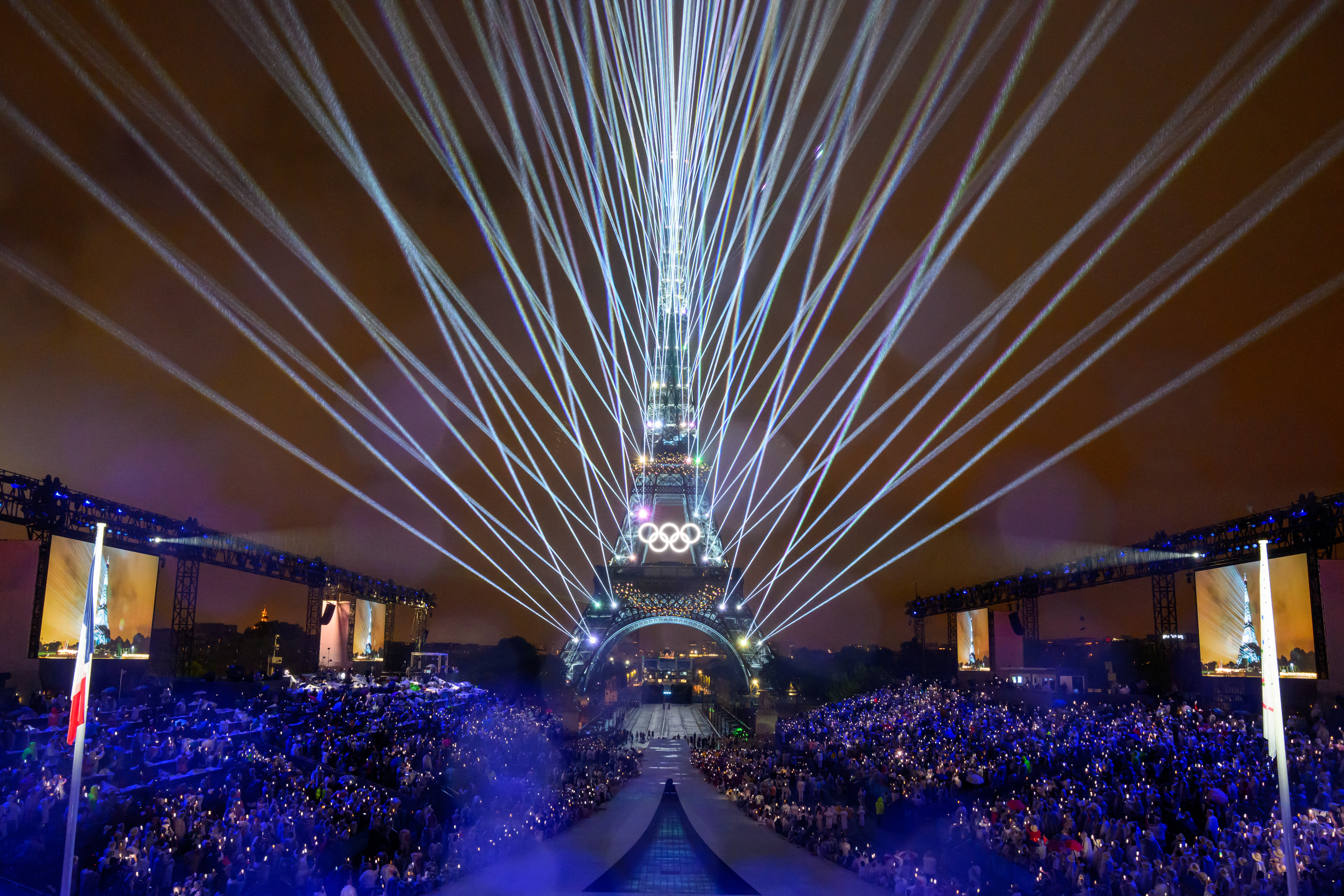 Lasers light up the sky around the Eiffel Tower, watched by a crowd at the Olympics opening ceremony.