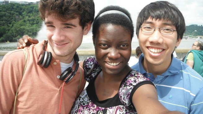 A white man, a Black woman, and an Asian man smile with their arms around each other in a sunny park
