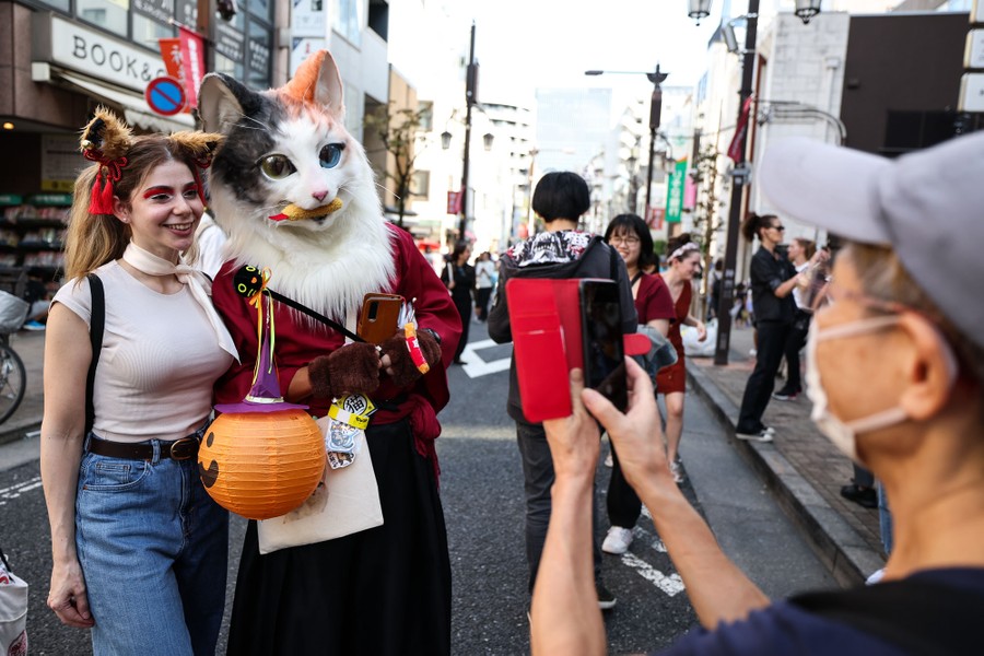 A person takes a photograph of two people, one wearing a full-head cat mask.