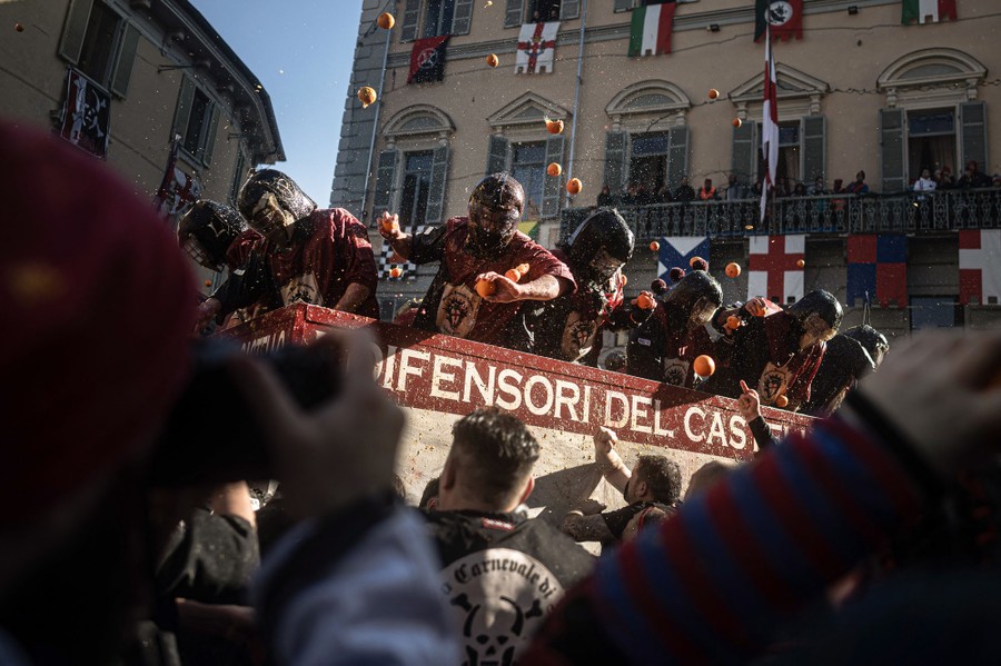 About a dozen people atop a truck, wearing protective helmets, throw oranges at a crowd below—who throw oranges back at them.