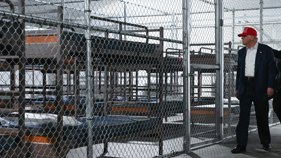 Photograph of Donald Trump in MAGA hat looking at rows of empty cots behind chain-link bars