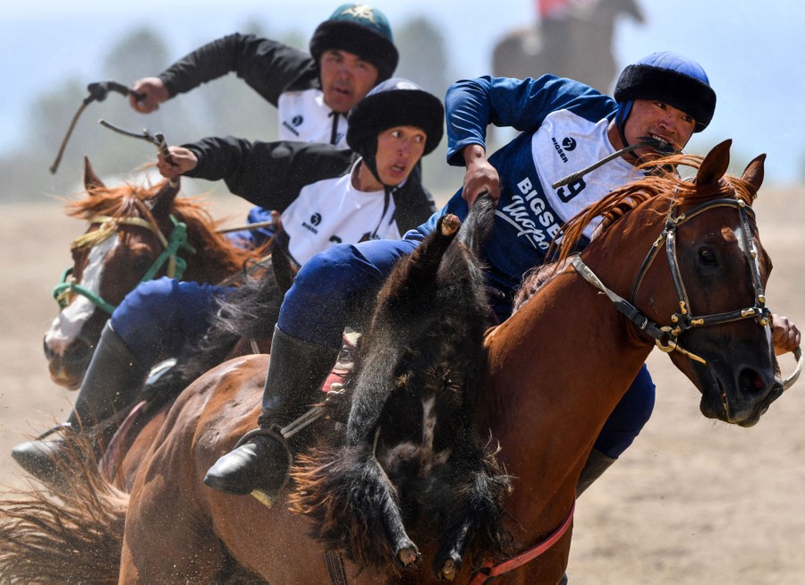 Three people ride horses at speed in a stadium while playing a game. The person in front holds a stuffed goat skin in their hand.