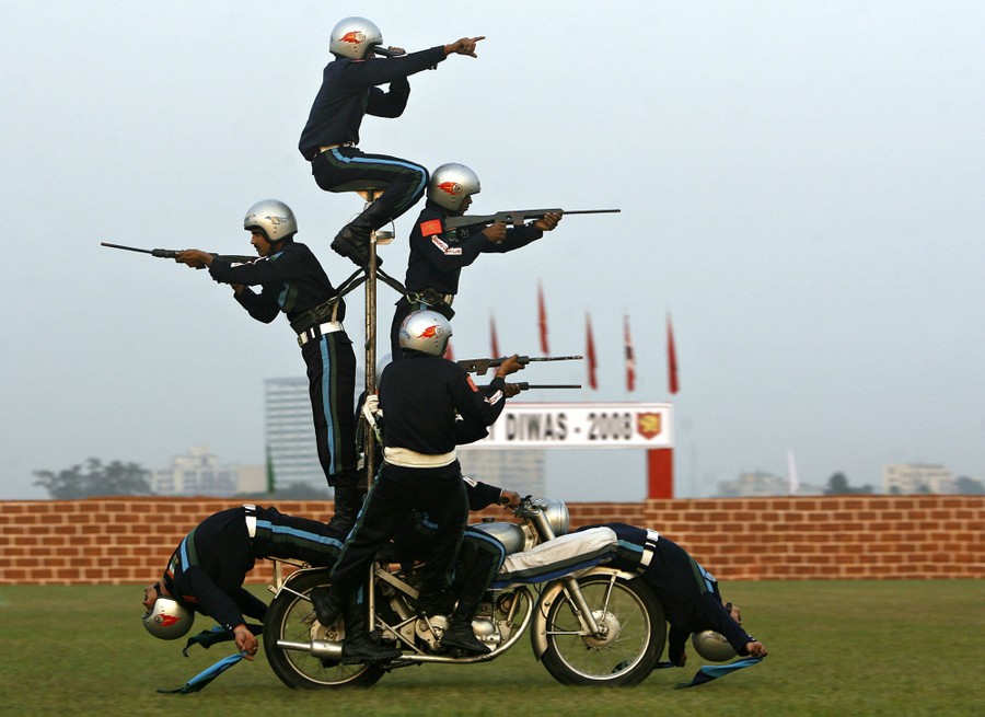 Eight stunt performers, four carrying weapons, ride on a motorcycle.