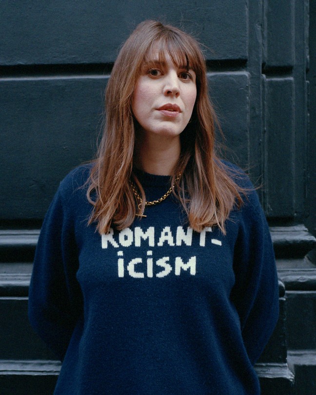 Alice Birch stands against a black wall with her hair blowing, wearing a blue sweater that reads "Romanticism."